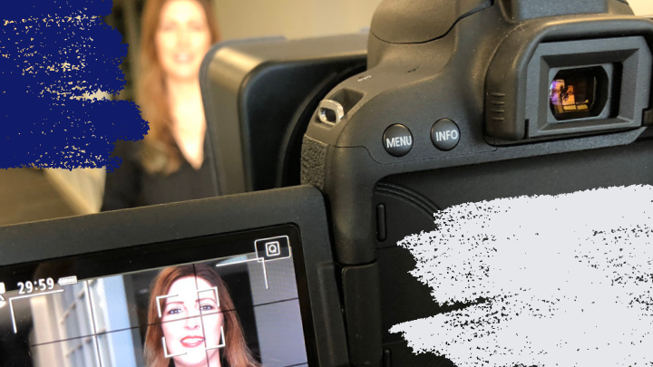 Looking from behind a camera pointed at a presenter with the presenter visible in the viewfinder screen